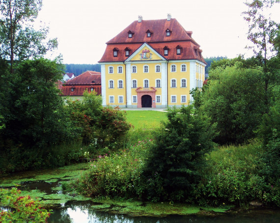 Das Foto zeigt das Schlossgebäude in Theuern, das in Mitten eines Parks mit Teich liegt.
